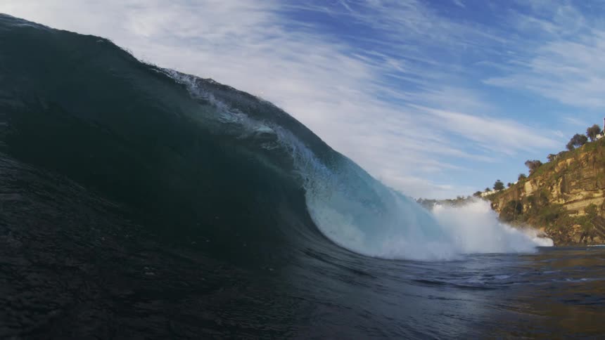 A point of view shot from the ocean shows a large blue wave crashing on the right with a surfer riding the wave in the distance and the top of a cliff with green trees and buildings visible in the background