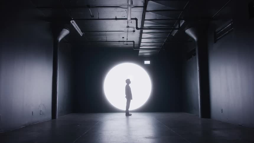 A man is standing still and silhouetted in a brightly lit circular spotlight against the dark backdrop of a large room