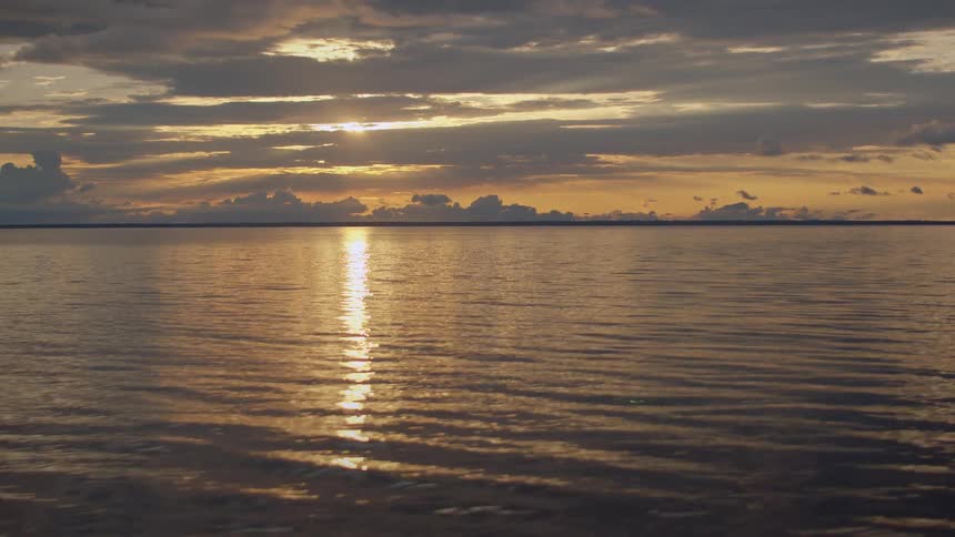As seen from a boat the golden light of the setting sun breaks through the clouds and reflects on the gentle ripples of the ocean