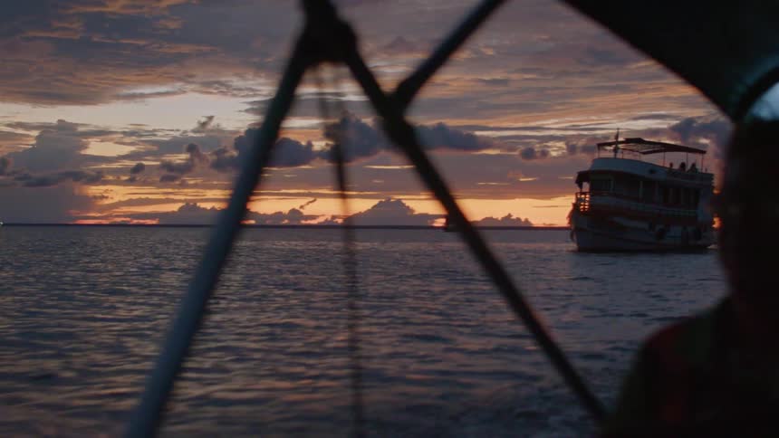 Man pilots his boat past a multi deck ship under a red tinted sky