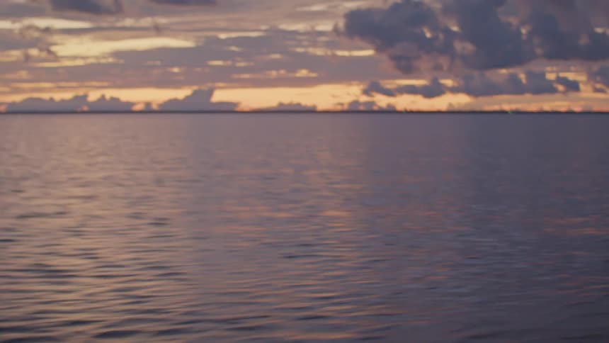 The camera pans up from the churning wake of a moving boat to reveal a beautiful wide landscape of the ocean under a dramatic sunset sky