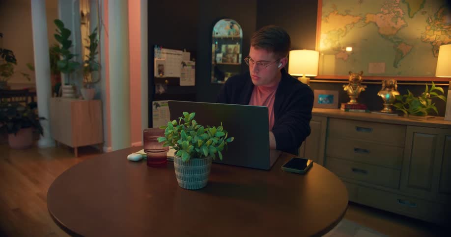 Man wearing glasses works on a laptop at a round table surrounded by plants and decorative items