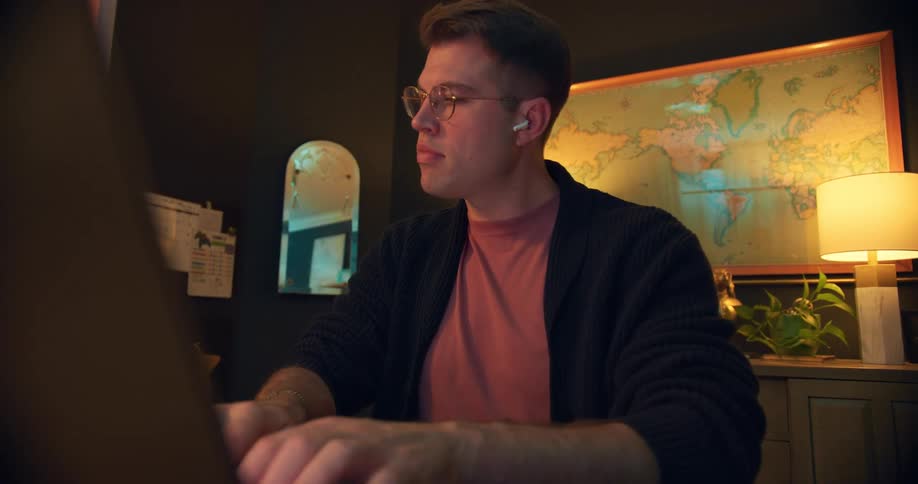 A young man wearing reading glasses is sitting indoors typing on a laptop