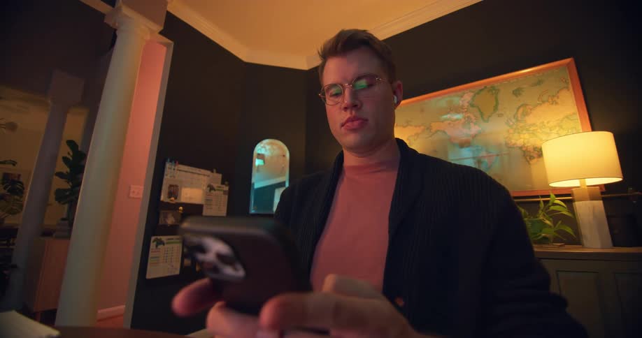 A man texts on his phone while sitting at a desk in a dim room
