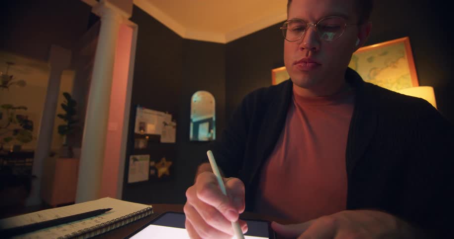 Man with glasses and earphones uses a stylus on a lit tablet
