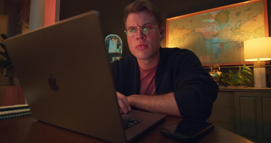 A man wearing glasses leans forward and looks at his laptop screen