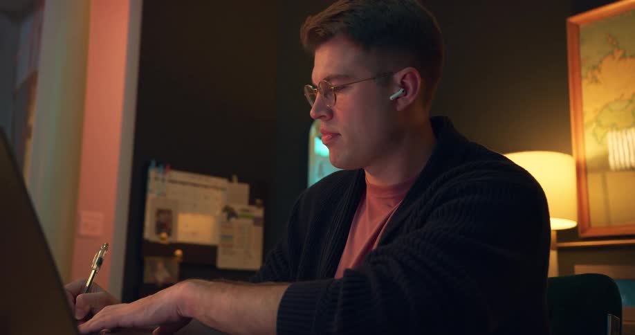 A focused young man with glasses and earbuds works at his desk late at night writing on a tablet before pausing to take a drink from his glass
