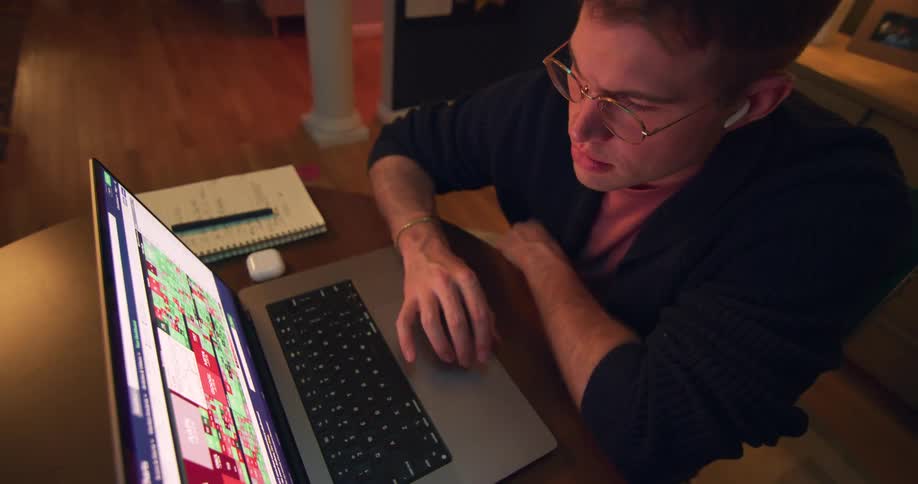 A focused man wearing glasses and earbuds is intently looking at a computer screen that displays a stock chart with red and green squares he is using the trackpad on his laptop and appears to be in an office or study environment