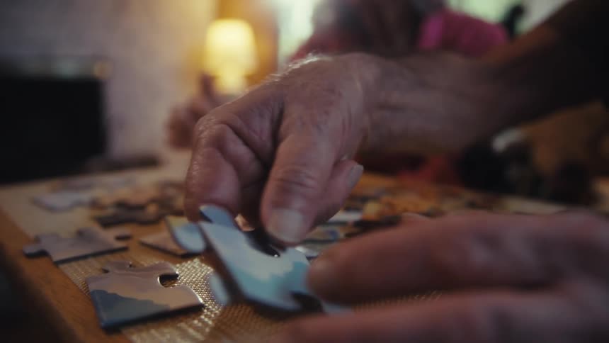 Close up of an elderly man and woman arranging a jigsaw puzzle on a table in a living room during daytime