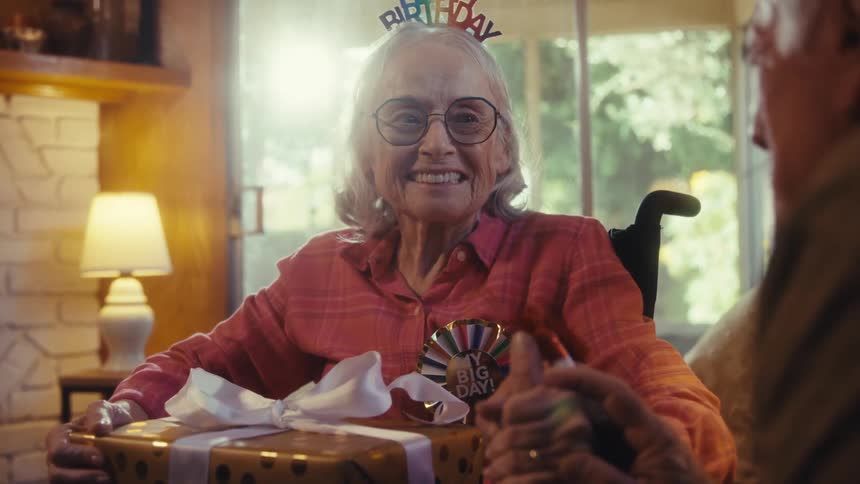 Elderly couple holding hands and smiling with a gift over the womans lap and a birthday hat over her head indoors during day time