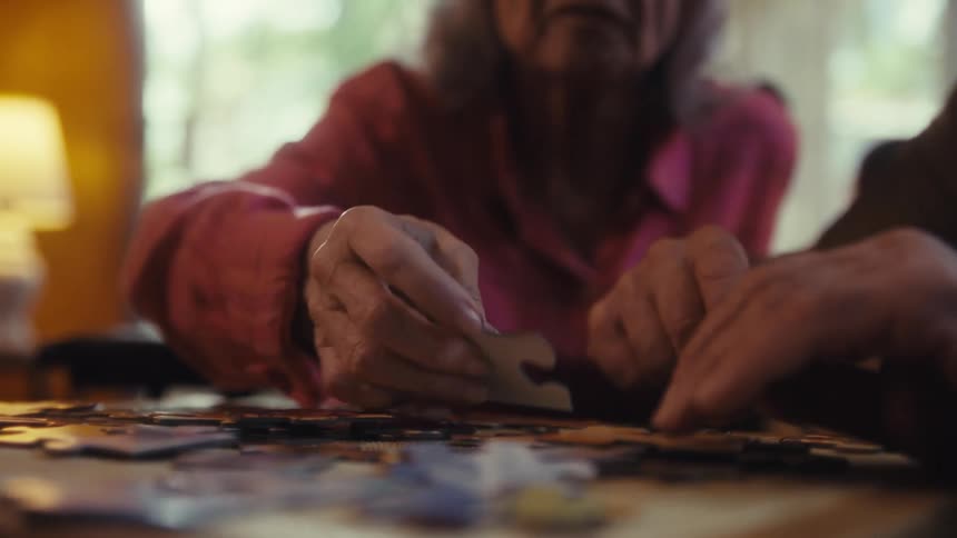 Close up of an elderly man and woman arranging a jigsaw puzzle on a table in a room during daytime