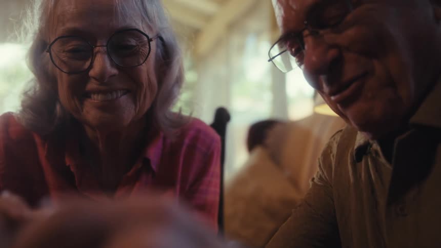 Two elderly people a woman and a man are sitting together at a table and working on a jigsaw puzzle they are focused on the pieces and the man gestures with his hand as he seems to be talking to the woman