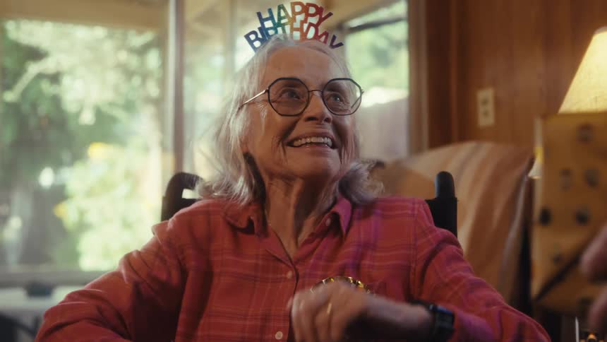 The video shows a closeup of an elderly woman who is celebrating her birthday she is wearing a happy birthday headband and a my big day ribbon pin she is first shown looking surprised and happy while holding a cell phone and then receives
