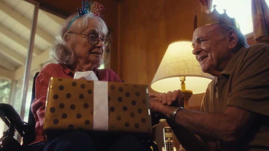 Elderly man shares smiles and conversation while gifting an elderly woman a present during the daytime at home