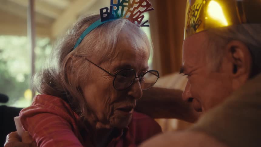 An elderly couple looking at each other as they talk and smile and the woman is wearing a hat with birthday writing on it