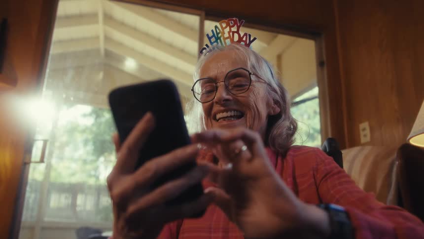 Smiling elderly woman with a happy birthday headband uses a phone