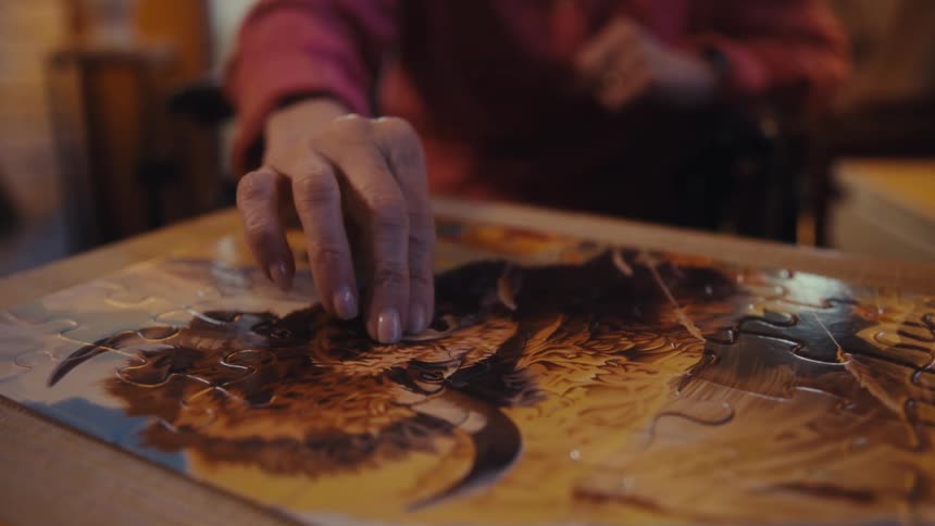 An elderly woman fits the last piece of a jigsaw puzzle on a table in a living room during daytime