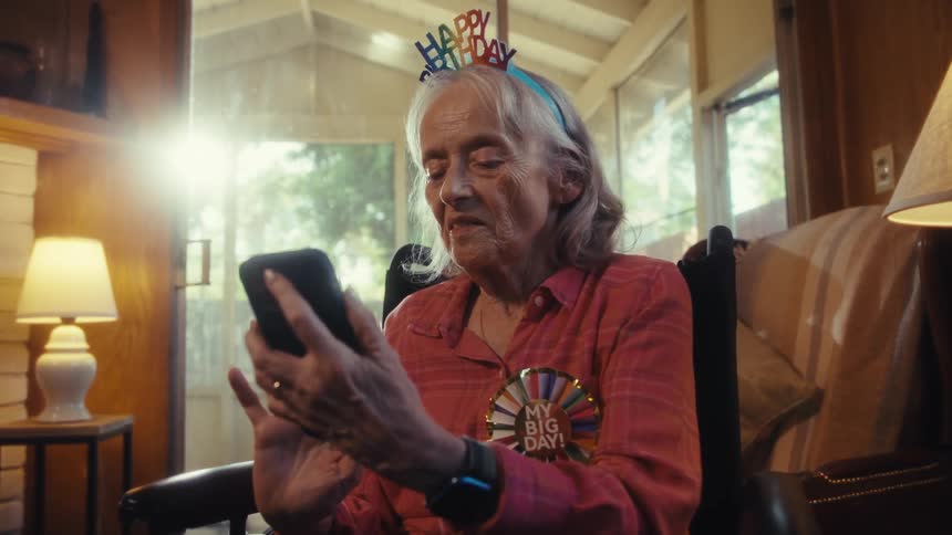 Elderly woman with a happy birthday headband holds a phone while smiling and talking