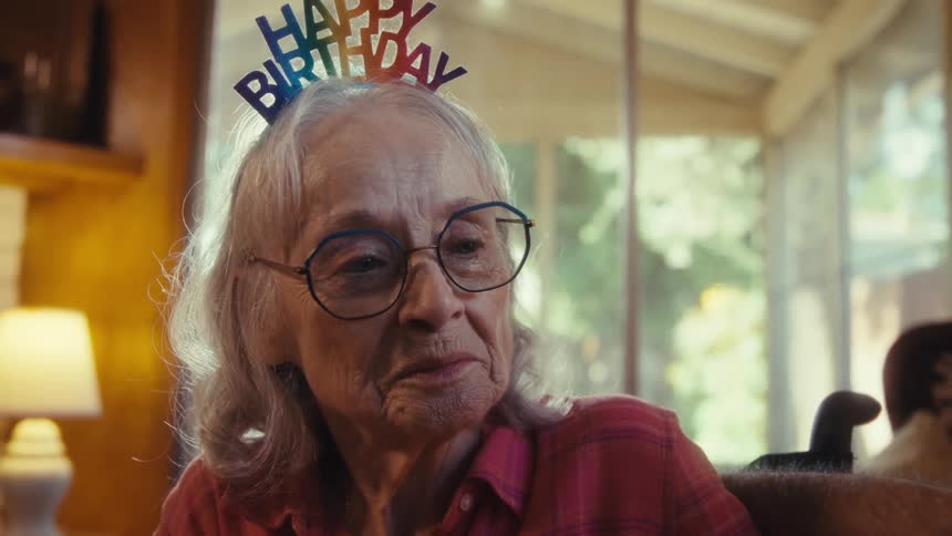 An elderly woman in a birthday headband sits thoughtful in warm afternoon light