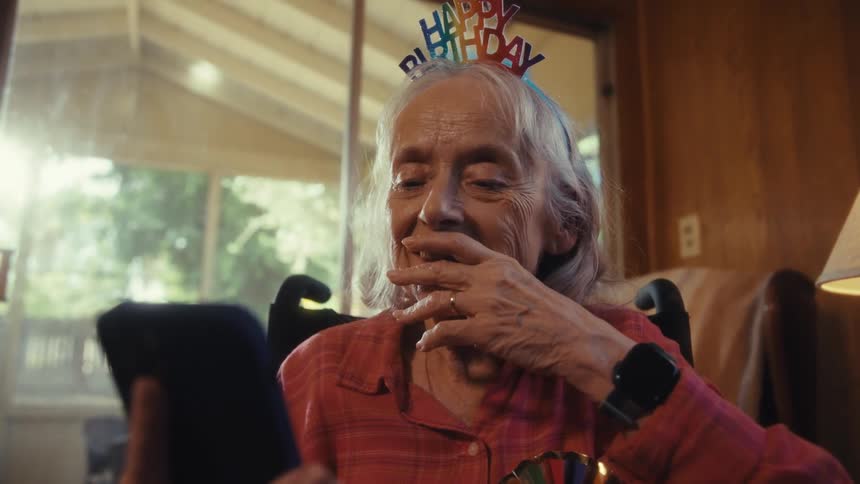 Smiling elderly woman with a happy birthday headband uses a phone on her special day