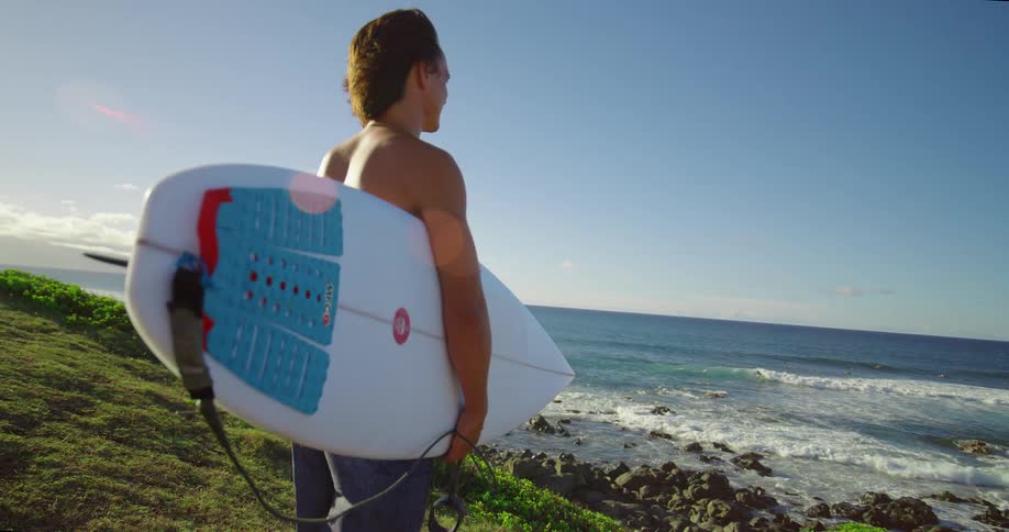 Young man stands on a hill overlooking the coast holding his surfboard and watching the waves roll into shore