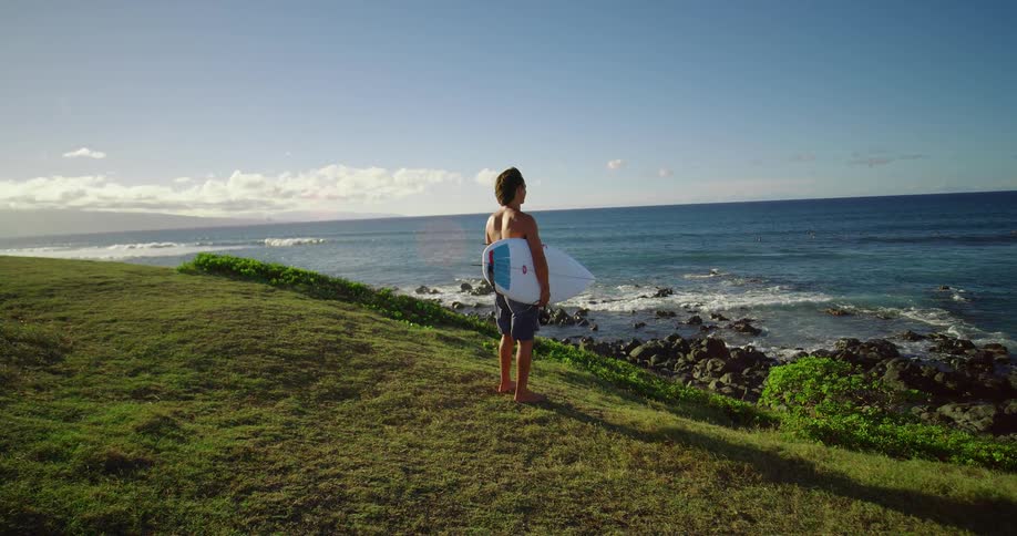 Men in shorts holds a board and looks at the sea from the cliff