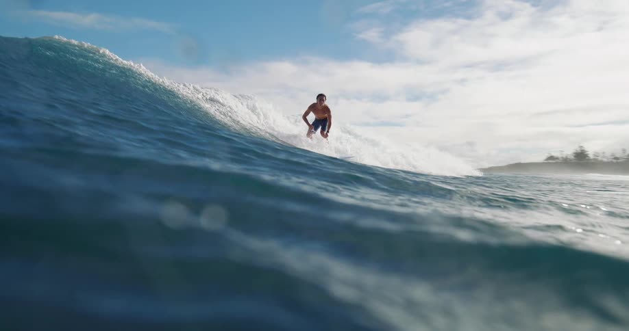 A surfer rides a wave under a bright blue sky with scattered clouds
