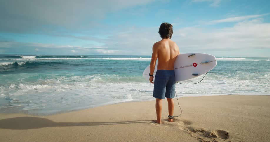 A person holding a surfboard is standing on the sandy beach looking out at the ocean waves ready to go surfing