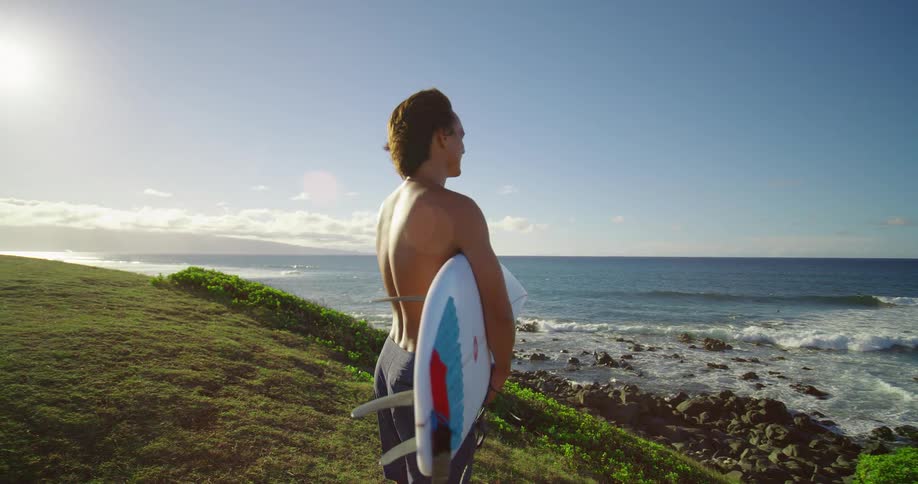 A man stands on a cliff with a surfboard under his arm looking out at the ocean