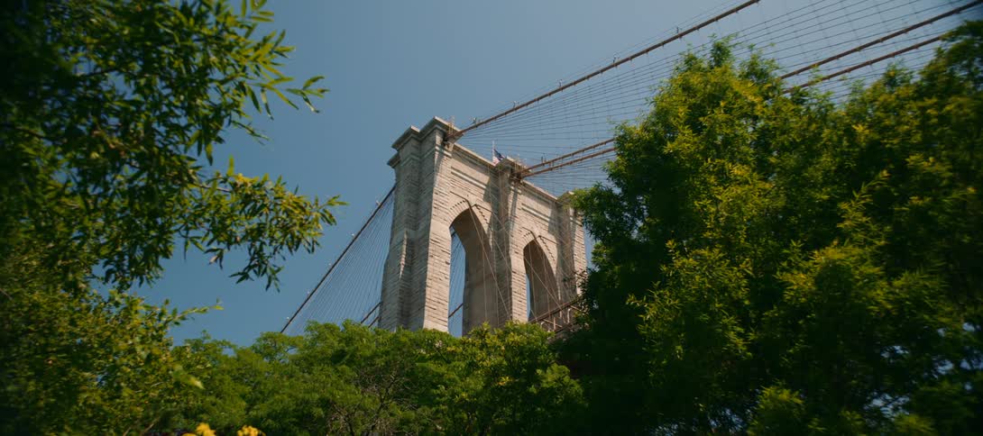 An upwardangle shot captures the iconic stone tower of the brooklyn bridge against a clear blue sky framed by lush green trees swaying gently in the breeze