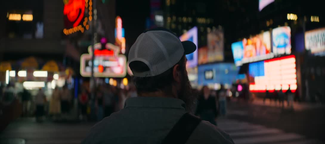 From behind a traveler stands in a glowing nightlife corridor absorbing the noise and color of giant signs