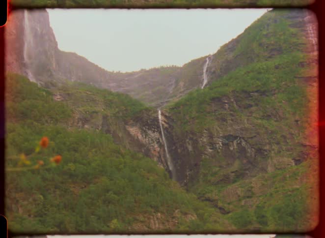 A rustic roofed shack stands below waterfalls on green mountains