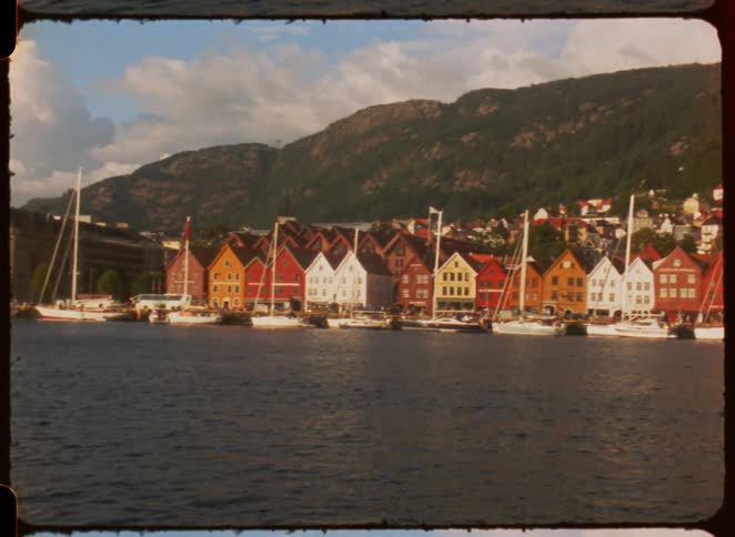 The video shot with a retro filter shows a scenic harbor with a row of colorful gabled wooden houses along the waterfront several boats are docked in front of the buildings with a forested mountain rising in the background