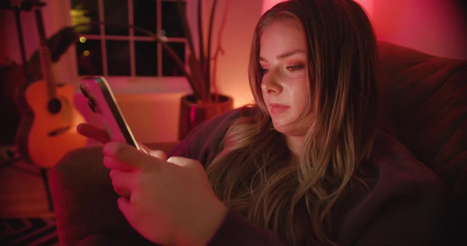 White woman sitting down on a couch using a smartphone indoors during night time