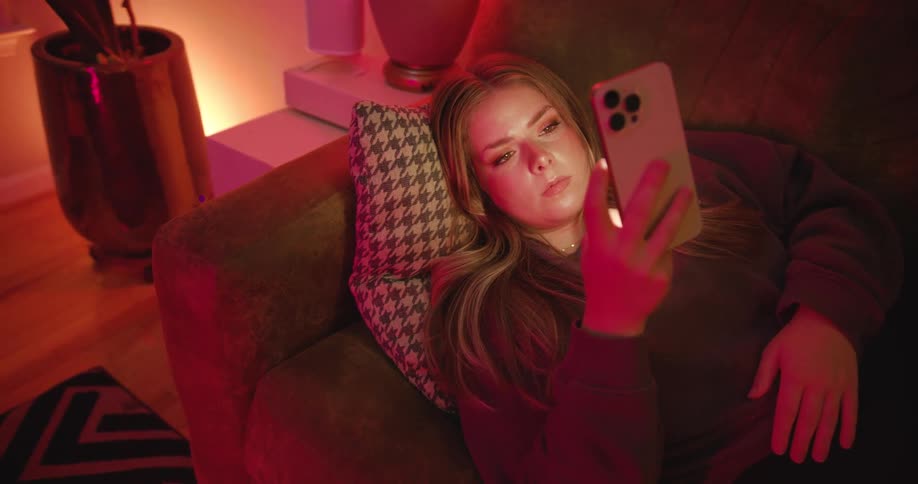 A young woman lays on her back on a couch and looks at her phone
