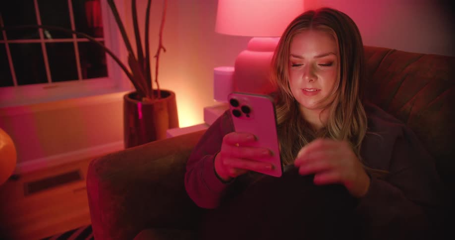 The video shows a young woman sitting on a couch in a dimly lit room illuminated by a soft reddishpink light she is engrossed in her cell phone holding it with both hands and scrolling with her thumb an acoustic guitar is visible in the backgroun