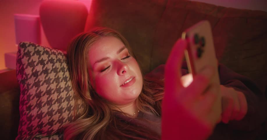 A woman laying on a couch smiles while using her smartphone