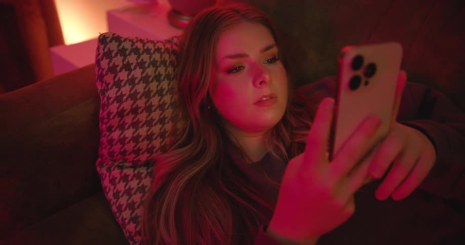 A woman lies on a couch looking at her phone under red lighting