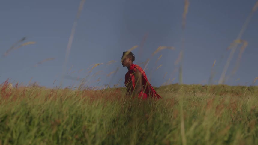 A man wearing a red outfit and is crouching in an environment with a lot of grass moving in the wind and he starts looking up