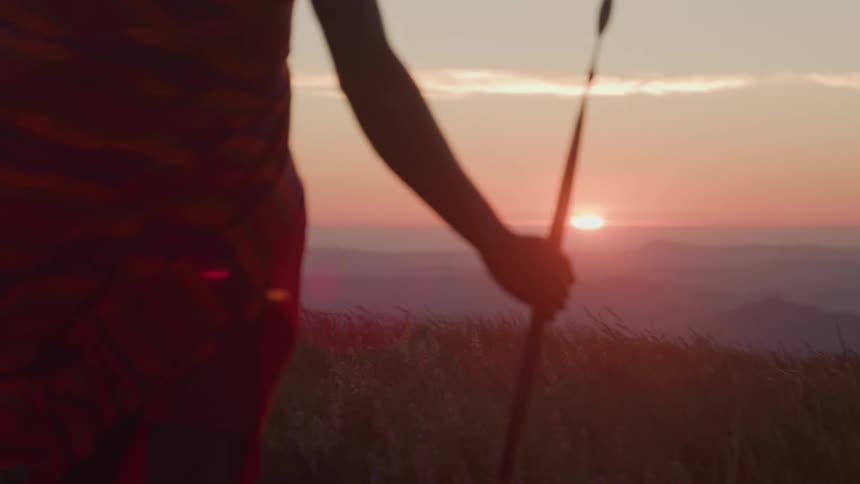 An african tribesman standing on a grassland wearing native attire and holding a spear watching the sunset