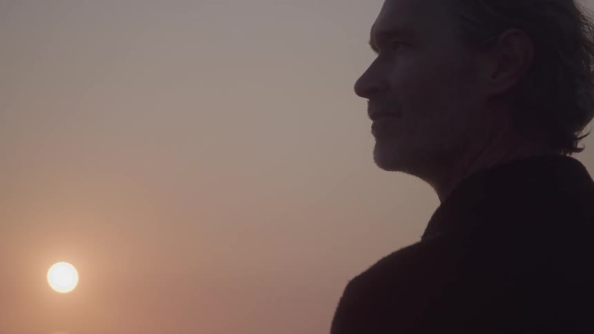 A man in profile with his hair tied back is silhouetted against a hazy sky the sun a bright circle is low on the horizon casting a soft warm glow