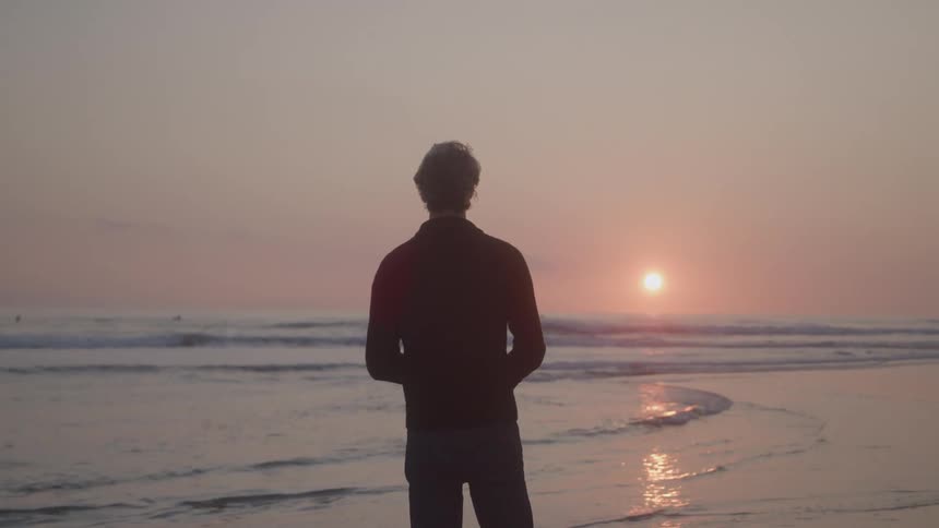 A man standing in front of a beach attacking some rocks in the sea water and the sun shining brightly in the morning