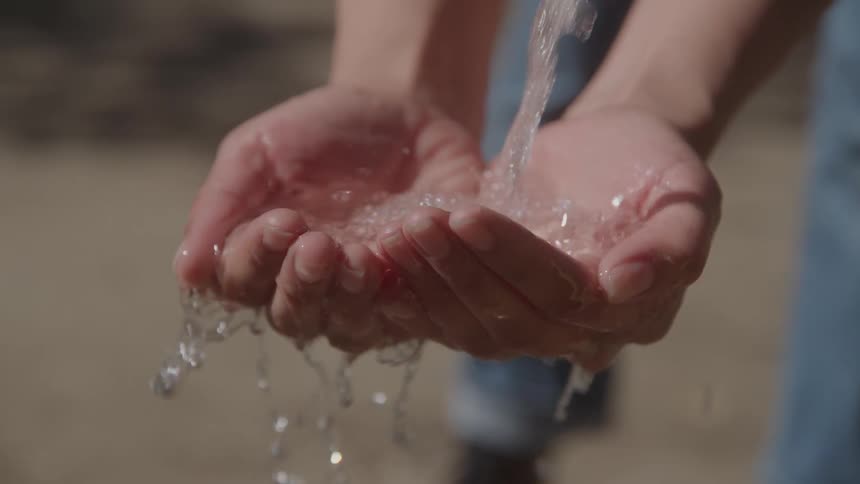 A closeup shot shows a persons cupped hands as fresh water pours into them then overflows and drips down the sunlight reflects off the glistening water making it appear clean and inviting