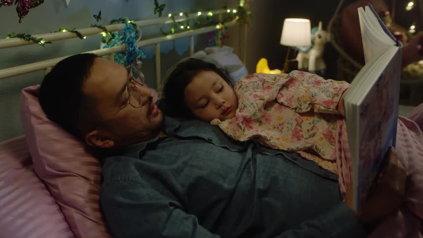 The video shows an intimate moment between a father and his young child they are lying in a cozy bedroom with pink blankets and string lights on the wall and the father is reading a book to the child who is nestled in his arms