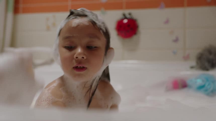 A mother wearing glasses bathing her daughter in a bathtub with a sponge while the girl has fun