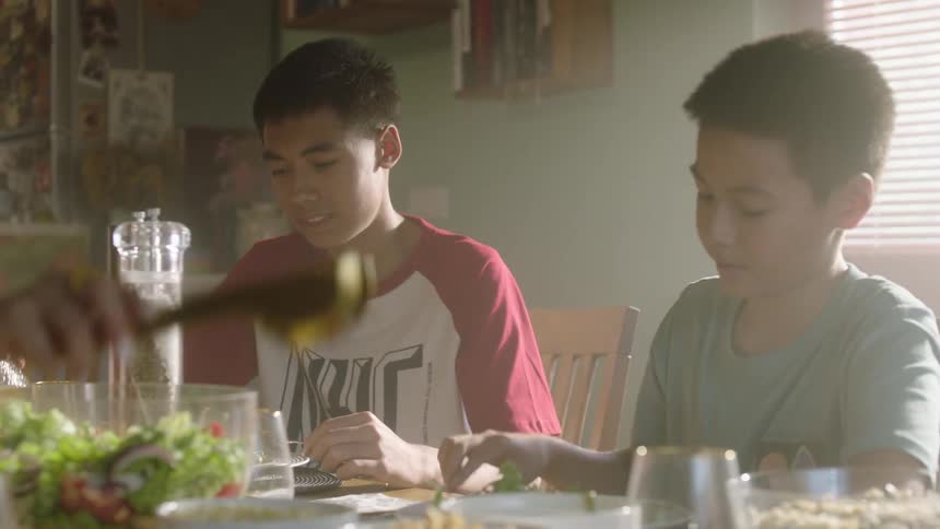 A family is eating a meal together at a dining table they are all seated and eating from bowls and plates and there are several dishes on the table
