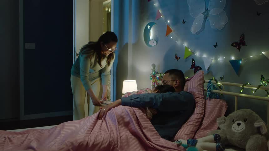 A father falls asleep while reading to his child and the mother quietly turns off the lamp and leaves the room