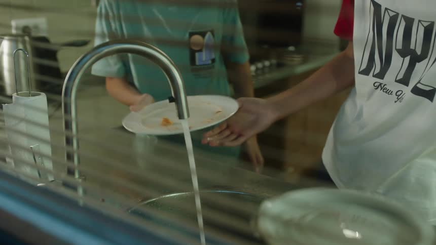 Two people are standing at a kitchen sink washing dishes they are using water from the faucet to rinse off plates before walking away