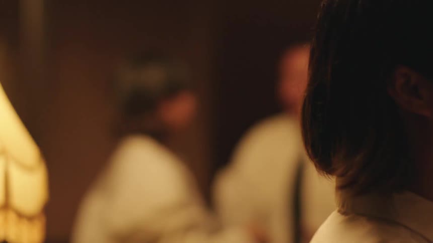 A couple is standing together in a dimly lit room with their faces close the woman looks at the man smiles and then turns her head away