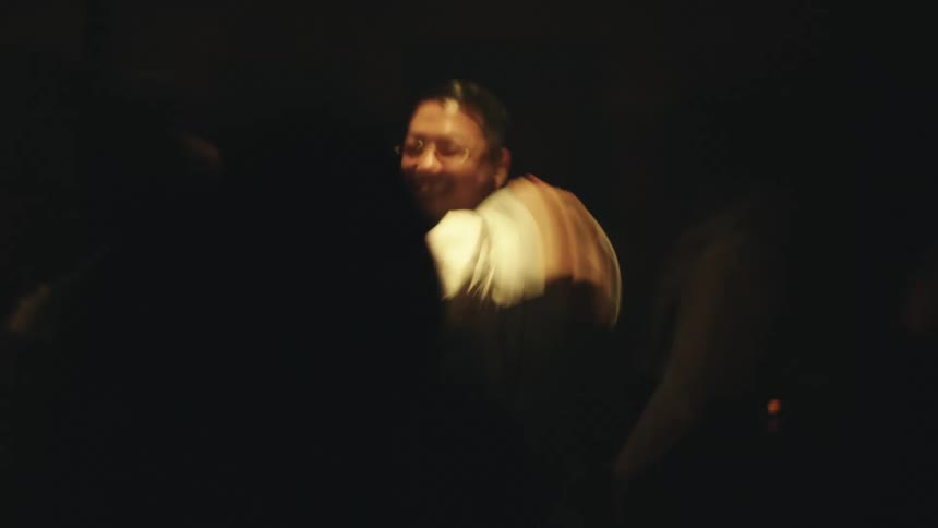 In a lowlight dark room a man in a brown suit and a woman in a white shirt are dancing on a timelapse scene